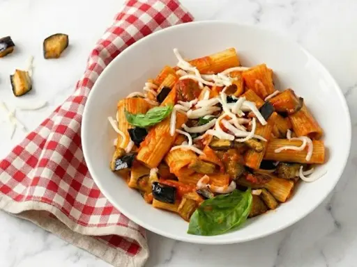 Un piatto di pasta alla norma fumante, con melanzane fritte, pomodoro e ricotta salata grattugiata, guarnito con basilico fresco.