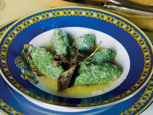 Strangolapreti alla trentina, verdi e saporiti, serviti in un piatto blu decorato con burro fuso e parmigiano.