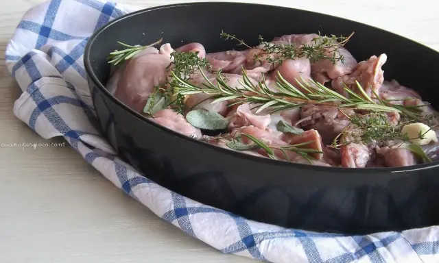Pezzi di coniglio crudo in una teglia, pronti per essere cucinati al forno con rosmarino, salvia e aglio.