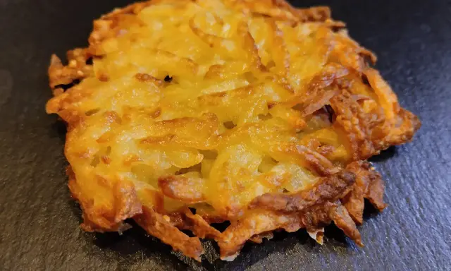 Un croccante rosti di patate dorato, con bordi leggermente bruciacchiati, adagiato su una superficie scura.