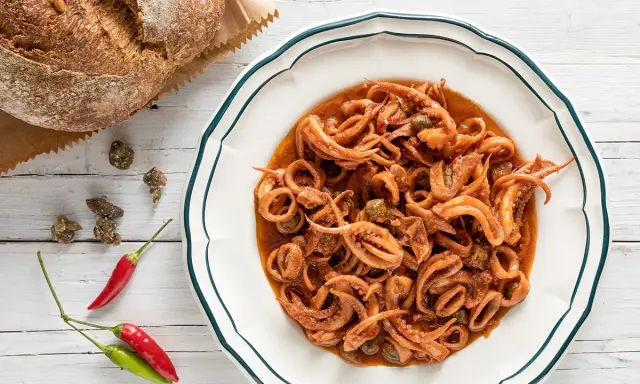 Un piatto invitante di totani in umido, con capperi e peperoncino, accanto a un pane rustico.