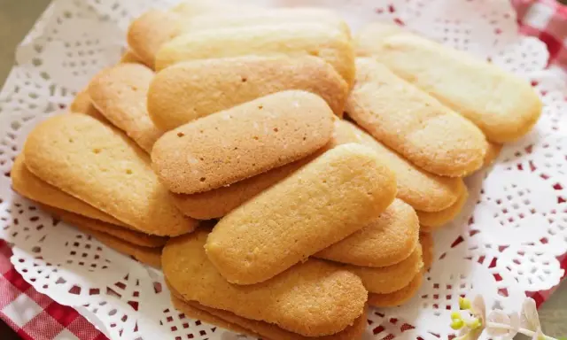 Un mucchio di biscotti a forma di lingue di gatto, dorati e friabili, disposti su una tovaglietta di pizzo bianco.
