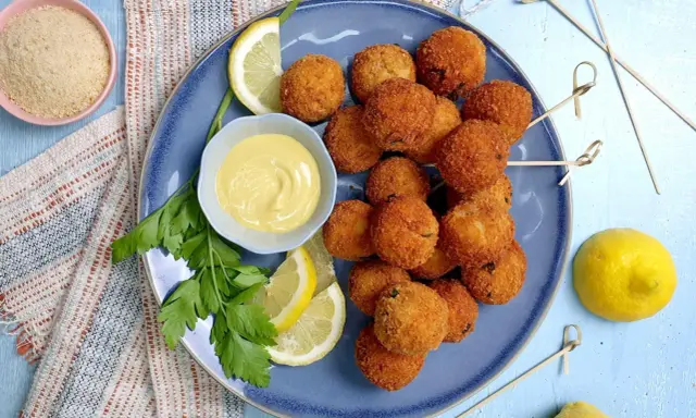 Piatto di polpette di tonno dorate e croccanti, servite con spicchi di limone, prezzemolo e una ciotolina di salsa gialla.