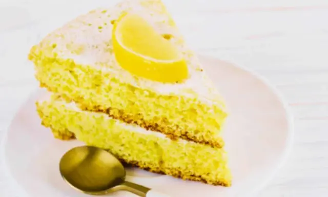 Fetta di soffice caprese al limone, spolverata di zucchero a velo e guarnita con una fetta di limone. Un cucchiaino dorato attende.
