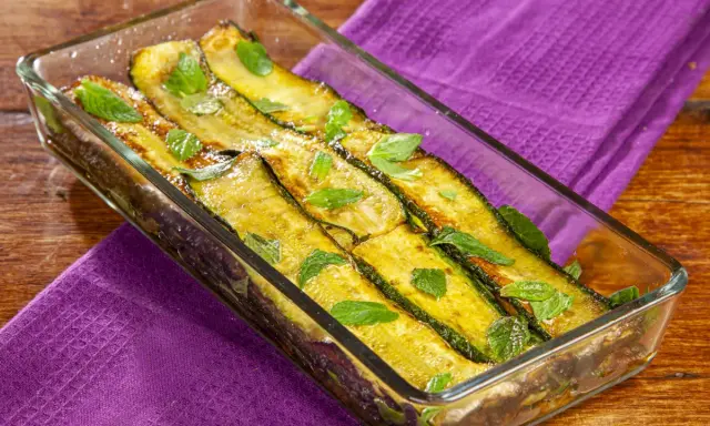 Zucchine alla scapece dorate e profumate alla menta, servite in una pirofila di vetro su un tovagliolo viola.