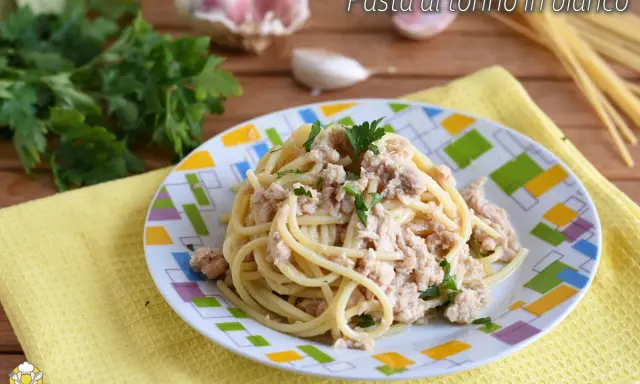 Un piatto di pasta al tonno in bianco, condita con tonno e prezzemolo, su un piatto colorato. Sullo sfondo, aglio e prezzemolo fresco.