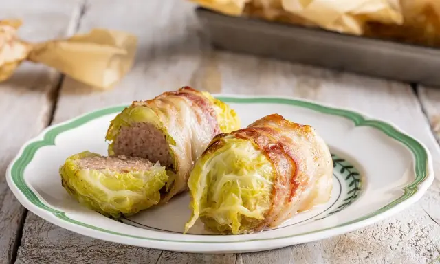 Deliziosi involtini di verza con ripieno di carne, avvolti in pancetta croccante, serviti su un piatto bianco con bordo verde.