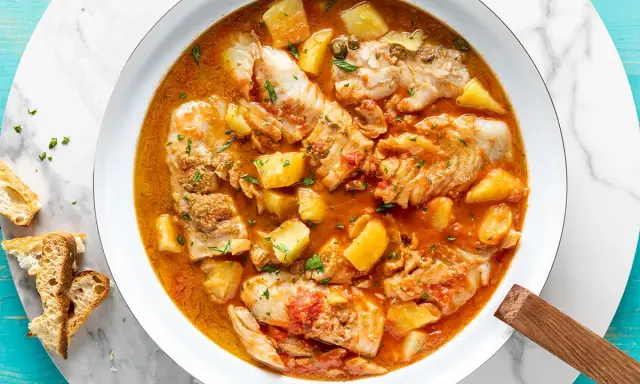 Un invitante merluzzo in umido con patate, pomodoro e capperi, guarnito con prezzemolo fresco e accompagnato da fette di pane croccante.