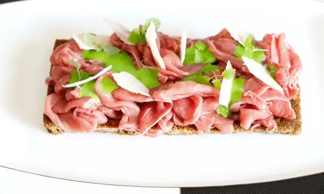 Delicato carpaccio di manzo su crostino integrale, guarnito con crema verde, scaglie di parmigiano e germogli freschi.