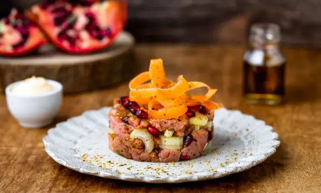Tartare di manzo con melograno e carote, servita su un piatto rustico. Sullo sfondo, spicchi di melograno e una bottiglietta di condimento.