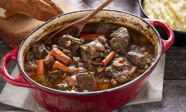 Un invitante spezzatino di manzo in un tegame rosso, con carote, funghi e un mestolo di legno. Accanto, pane rustico e purè di patate.