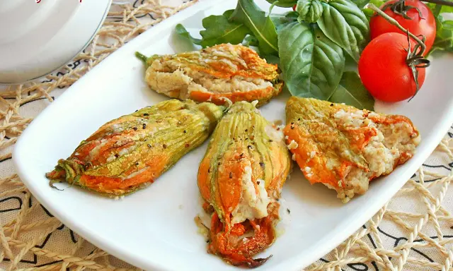 Deliziosi fiori di zucca al forno ripieni, serviti su un piatto bianco con pomodorini e basilico fresco.