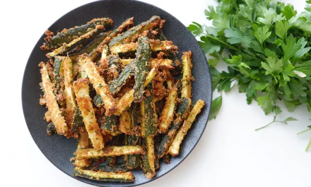 Zucchine gratinate in padella, dorate e croccanti, servite su un piatto nero con un ciuffo di prezzemolo fresco.