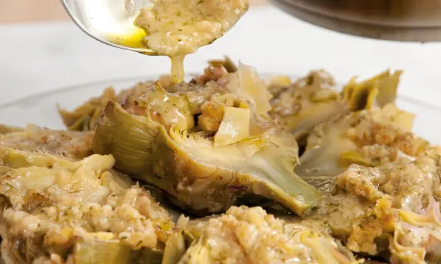 Un cucchiaio versa un sugo dorato su un piatto di carciofi alla romana, conditi con erbe aromatiche e pane croccante.