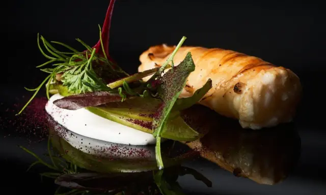 Spiedini di pesce avvolti in pasta sfoglia, serviti con una crema bianca e insalata fresca, su sfondo nero.