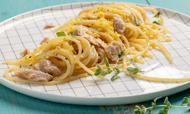 Un piatto di pasta tonno e limone, condita con scorza di limone grattugiata e erbe aromatiche fresche, servita su un piatto a scacchi bianchi.