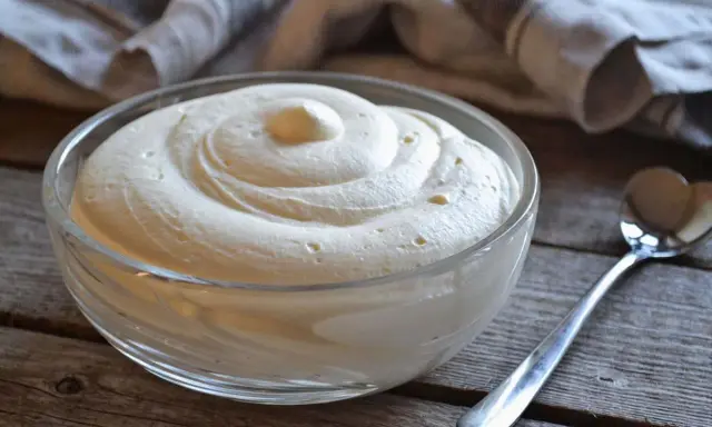 Una ciotola di vetro piena di soffice crema al mascarpone, con una spirale perfetta e una cucchiaio d'argento accanto.