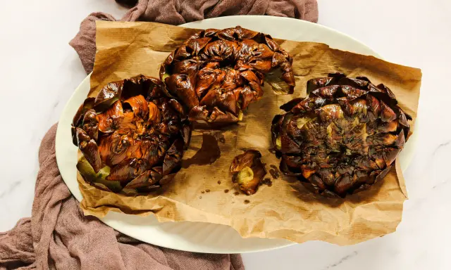 Tre carciofi alla giudia dorati e croccanti, serviti su carta da forno marrone, su un piatto bianco.
