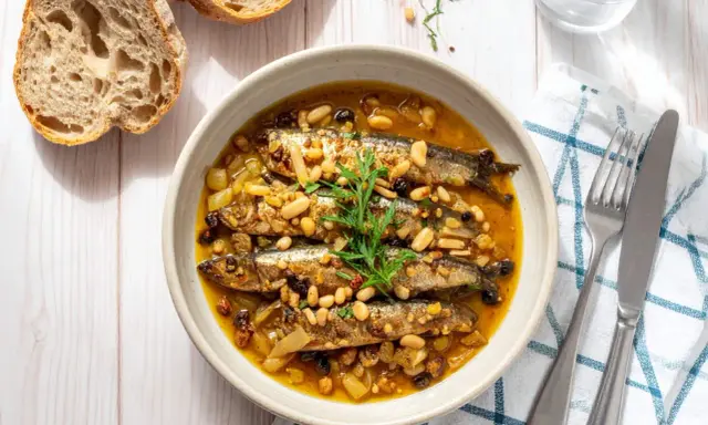 Sarde in saor in una ciotola, con pinoli, uvetta e cipolle. Accanto, pane rustico e posate.