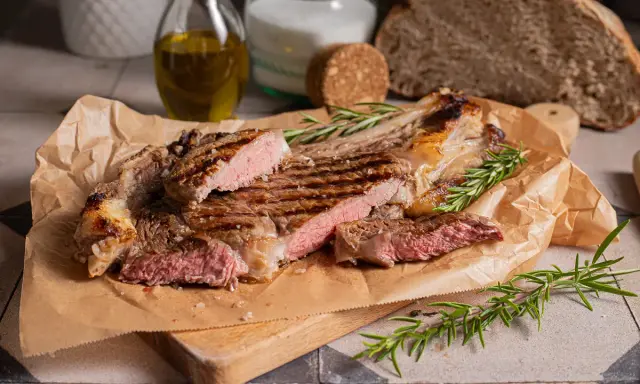 Succulenta costata di manzo alla griglia, tagliata a fette, con rosmarino fresco e sale grosso. Sullo sfondo, pane rustico e olio d'oliva.