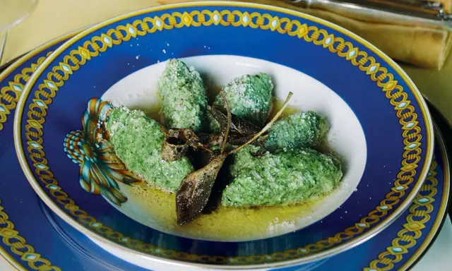 Strangolapreti alla trentina, verdi e saporiti, serviti in un piatto blu decorato con burro fuso e parmigiano.