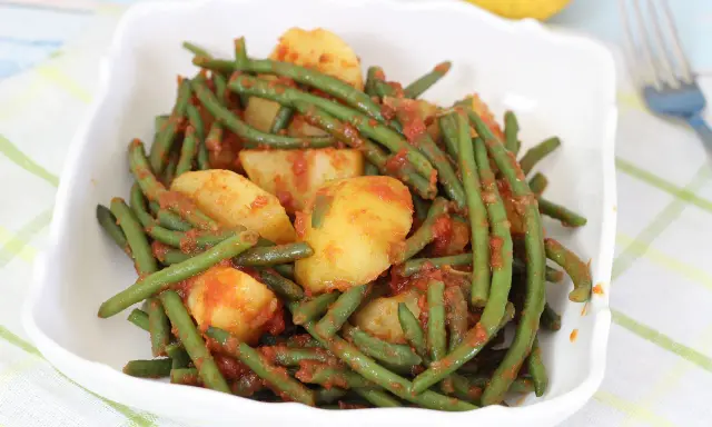 Un piatto di fagiolini in umido con patate, conditi con un sugo rosso saporito, servito in una ciotola bianca.