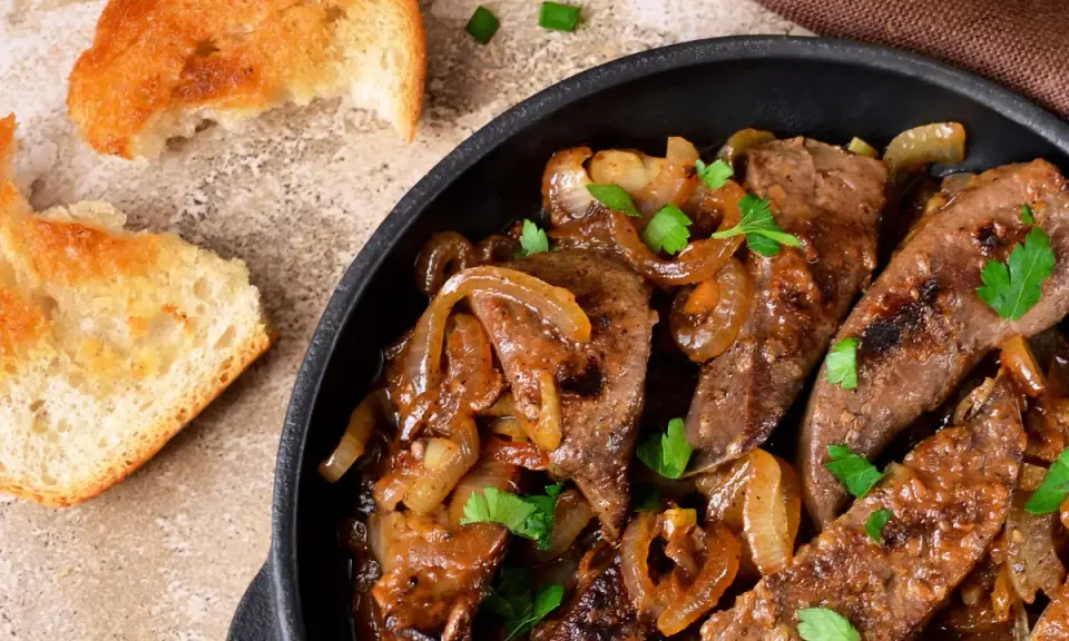 Fegato alla veneziana succulento in padella con cipolle caramellate e prezzemolo fresco, accompagnato da fette di pane tostato.