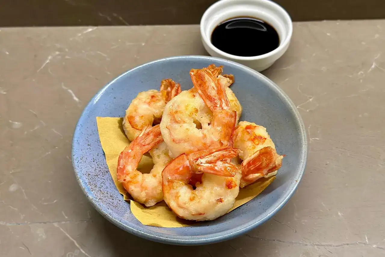 Gamberi in tempura dorati e croccanti, serviti su carta gialla in una ciotola blu, con una ciotolina di salsa scura sullo sfondo.