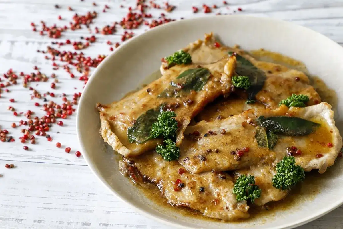 Scaloppine al vino bianco su un piatto, guarnite con salvia e prezzemolo, con pepe rosa sparso sullo sfondo.
