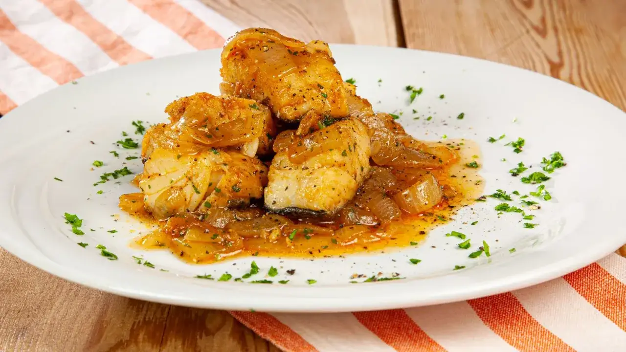 Pezzi di merluzzo in umido con cipolle dorate, guarniti con prezzemolo tritato e pepe nero su un piatto bianco.