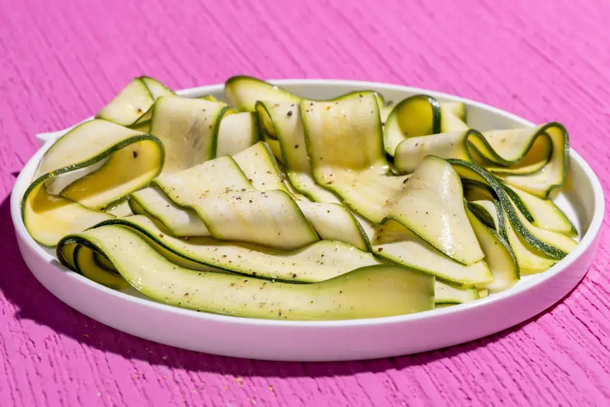 Fettine sottili di zucchine marinate crude, condite con pepe nero, su un piatto bianco. Freschezza estiva in ogni morso.