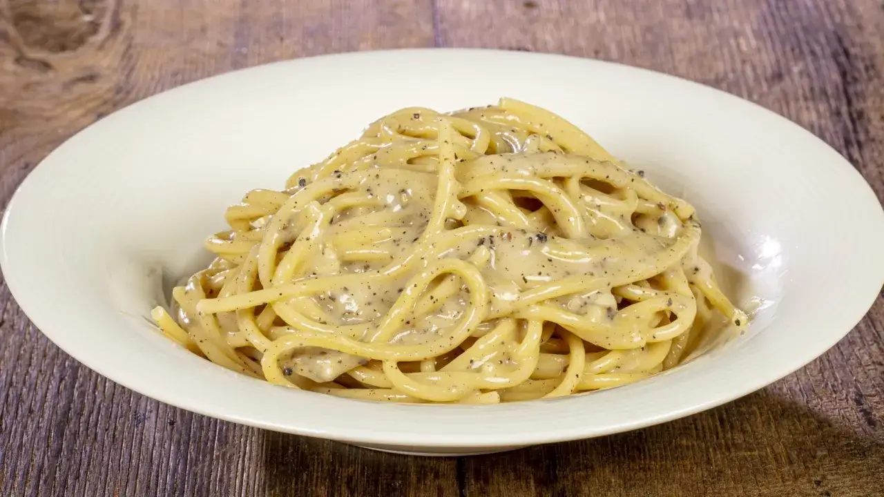 Un piatto di spaghetti cacio e pepe cremosi, conditi con abbondante pepe nero macinato, serviti in una ciotola bianca su un tavolo di legno.