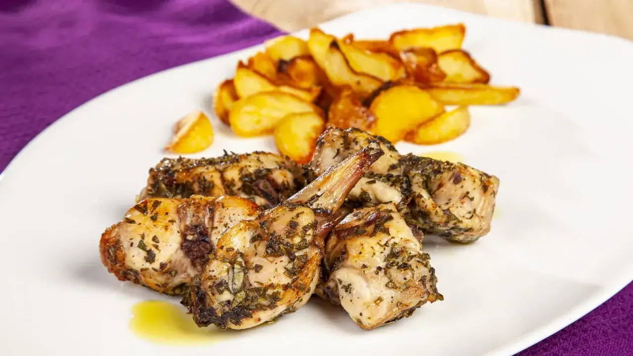 Pezzi di coniglio al forno, dorati e profumati alle erbe, serviti con patate arrosto su un piatto bianco.