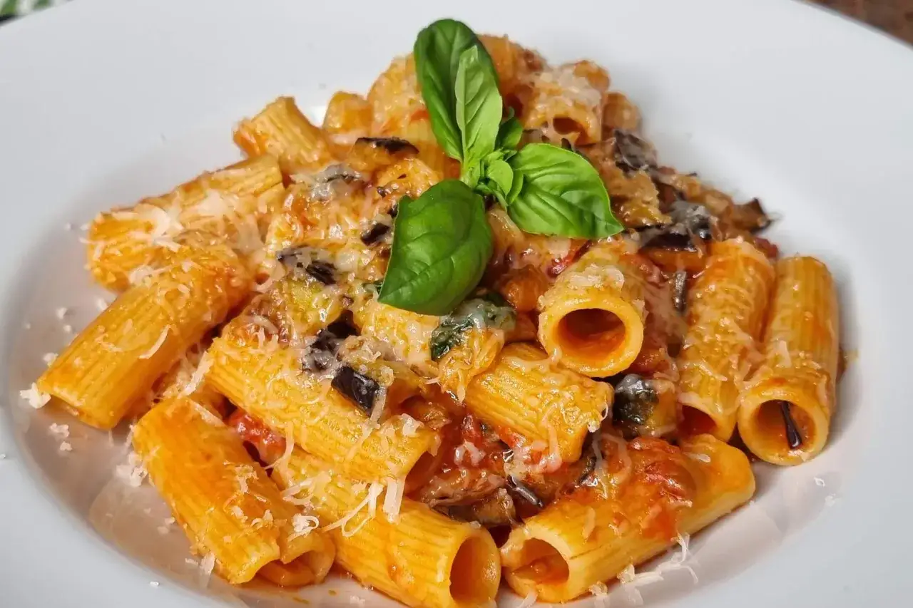 Un piatto di pasta alla norma fumante, con rigatoni conditi con melanzane, pomodoro e ricotta salata, guarnito con basilico fresco.