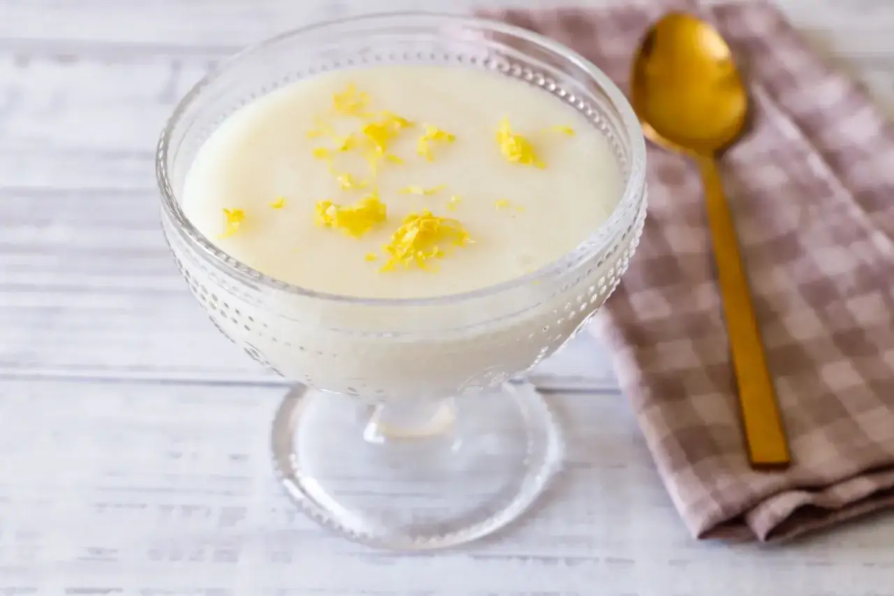 Una coppa di crema di riso guarnita con scorza di limone, accanto a un cucchiaio dorato e un tovagliolo a quadretti.