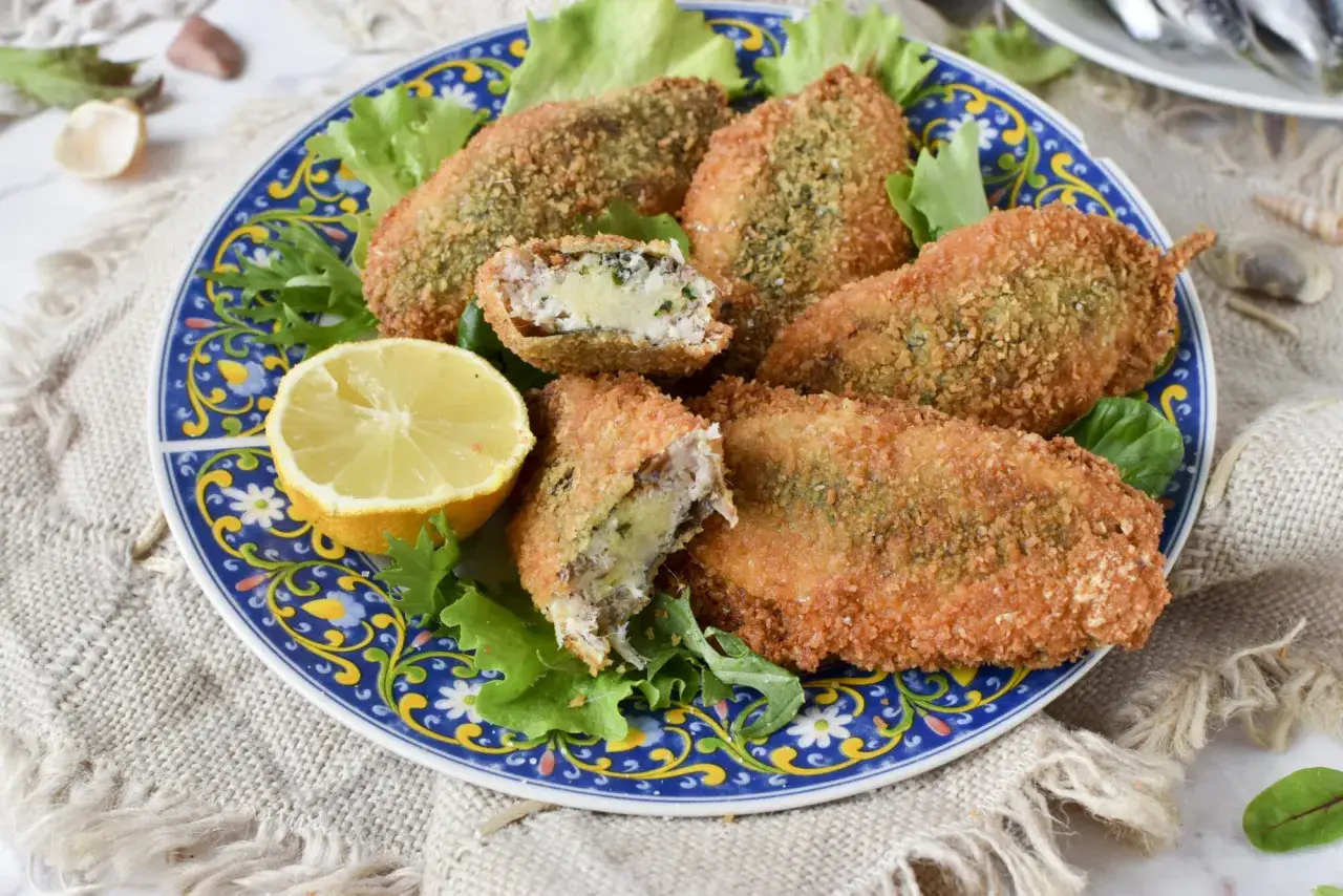 Sarde a beccafico dorate e croccanti, ripiene di un saporito impasto, servite su un piatto blu decorato con lattuga e spicchio di limone.