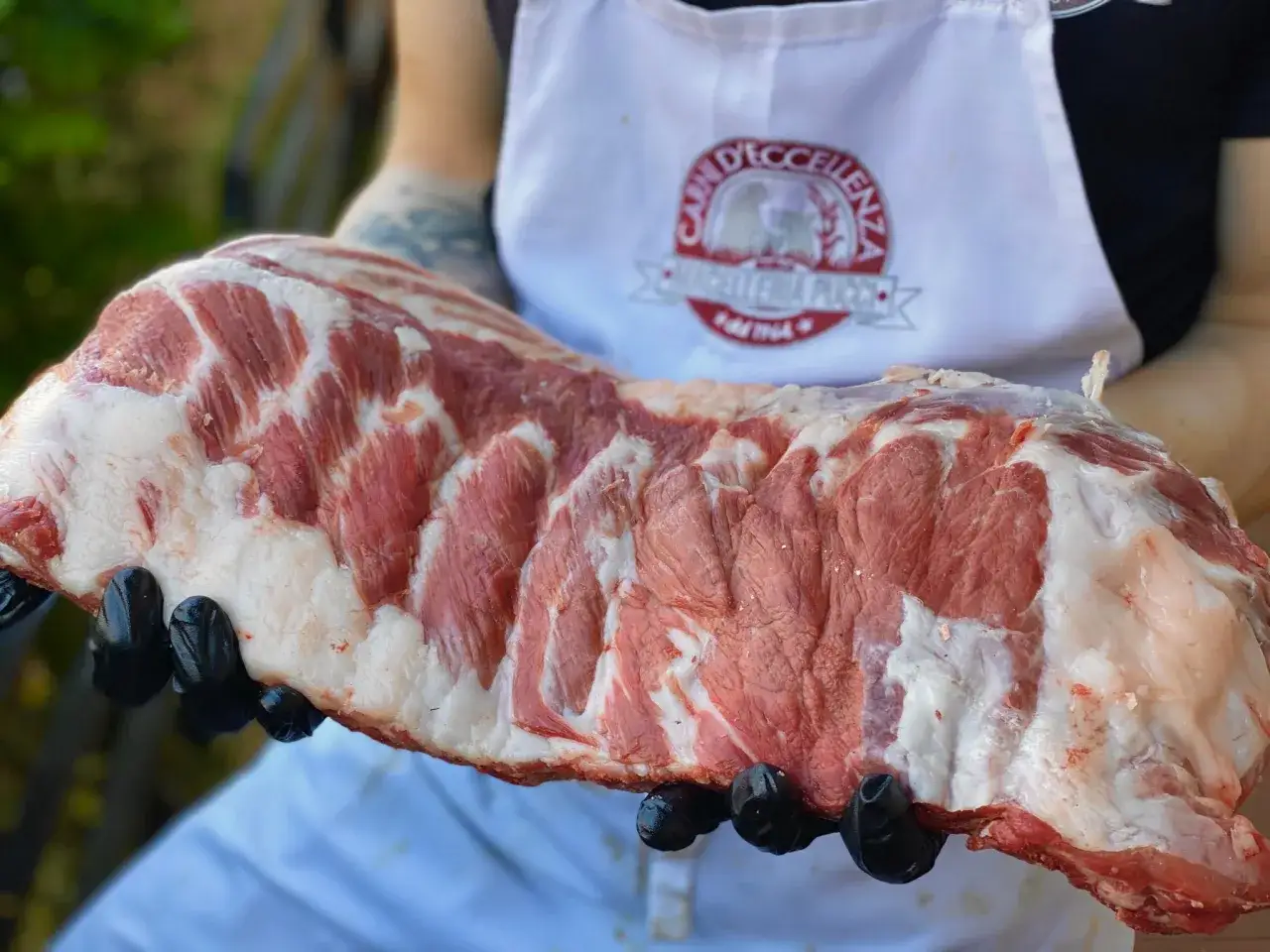 Una succulenta braciola di maiale, con striature di grasso bianco e carne rossa, tenuta in mano con guanti neri.