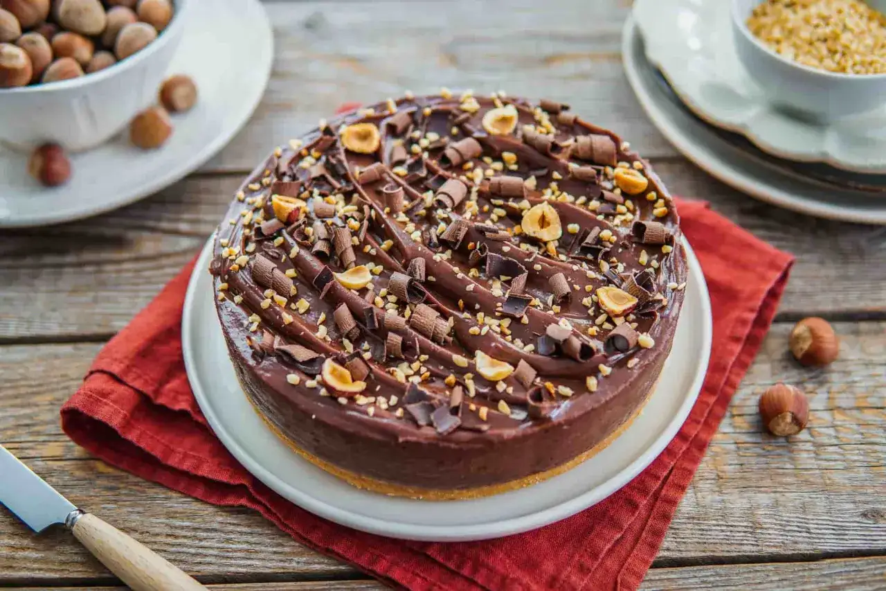 Cheesecake alla Nutella ricoperta di riccioli di cioccolato e nocciole tostate. Un sogno goloso su un piatto bianco, pronto per essere gustato.