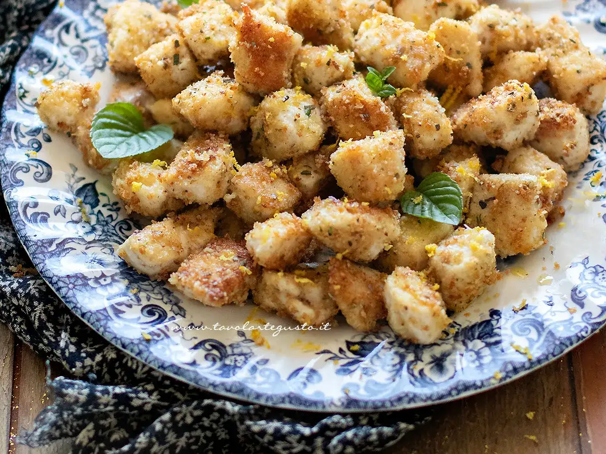Bocconcini di pollo dorati e croccanti, guarniti con scorza di limone e foglie di menta, serviti su un piatto decorato.