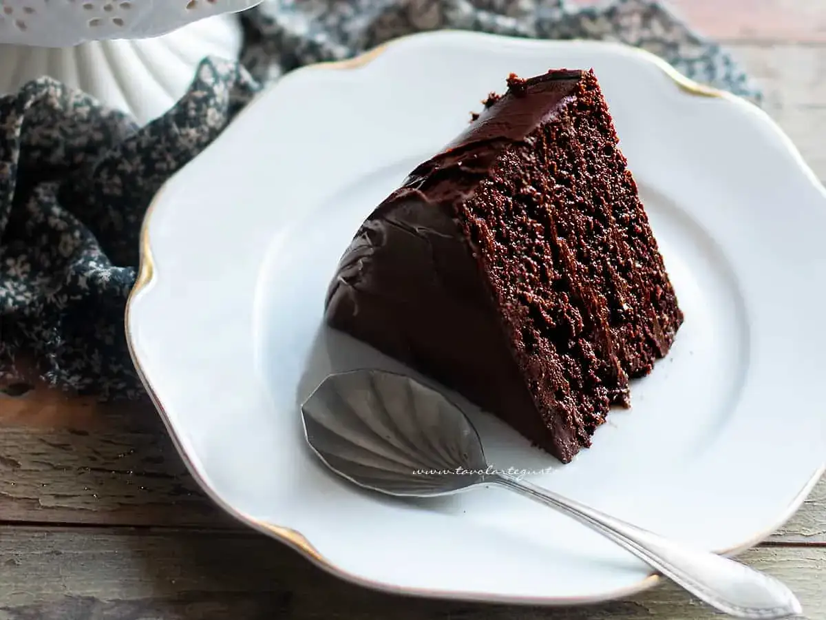 Una fetta di torta al cioccolato fondente, lucida e invitante, su un piatto bianco con bordo dorato. Un cucchiaino argentato attende accanto.