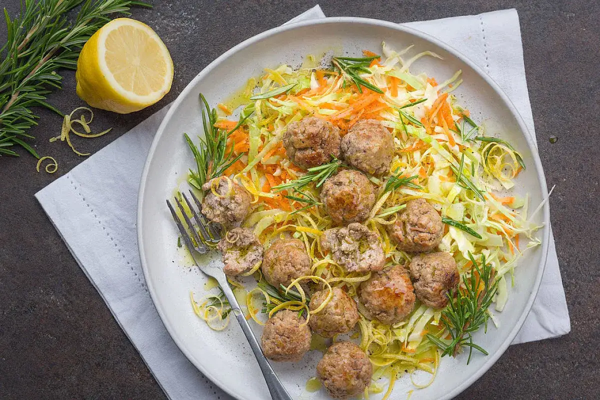 Polpette di carne morbide servite su un letto di insalata di cavolo e carote, guarnite con rosmarino e scorza di limone.