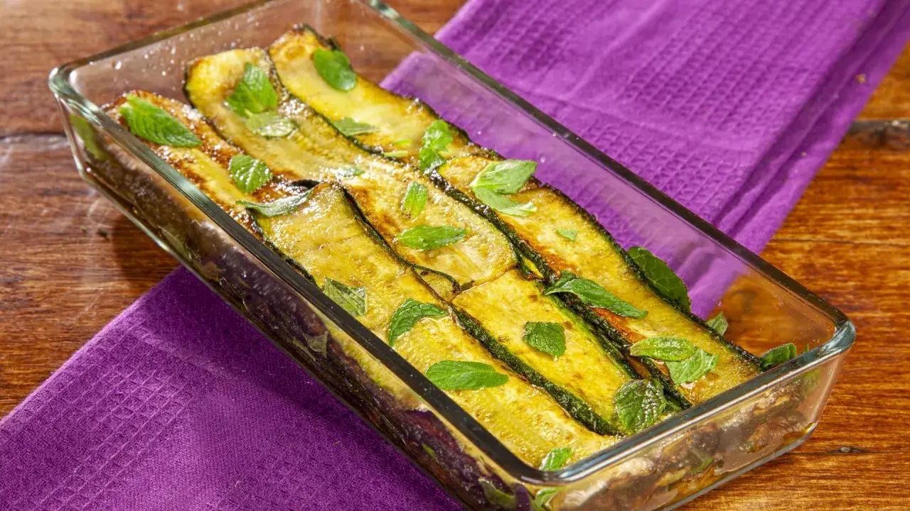 Zucchine alla scapece dorate e profumate alla menta, servite in una pirofila di vetro su un tovagliolo viola.