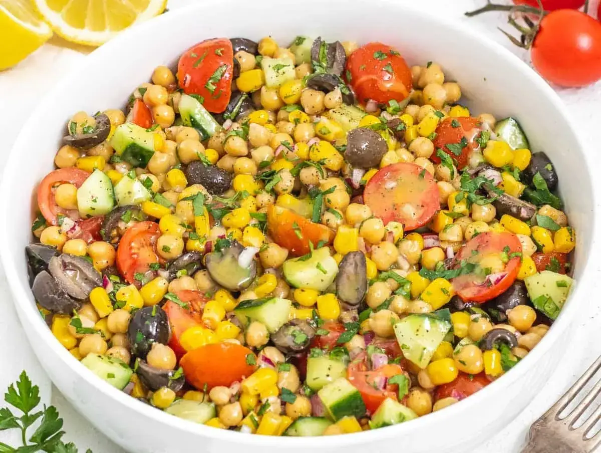 Fresca insalata di ceci con pomodorini, mais, olive nere e cetrioli, guarnita con prezzemolo tritato. Un piatto colorato e salutare.