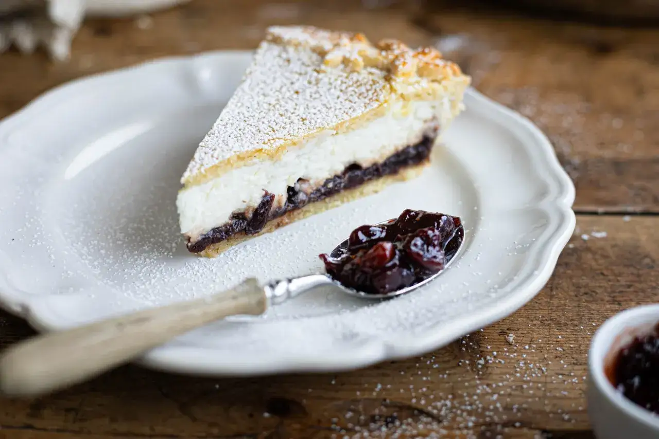 Una fetta di crostata ricotta e visciole, cosparsa di zucchero a velo, su un piatto bianco. Un cucchiaino &egrave; pieno di visciole sciroppate.