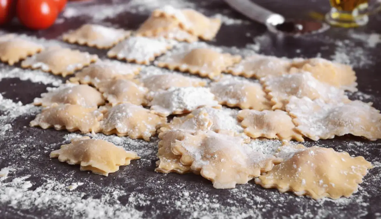 Tanti ravioli fatti in casa, spolverati di farina, pronti per essere cucinati. Sullo sfondo pomodori e un bicchiere d'olio.