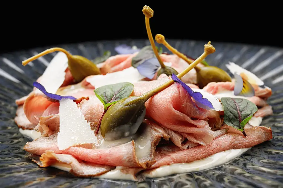 Vitello tonnato ricetta originale piemontese: fettine sottili di arrosto di vitello, salsa tonnata cremosa, capperi, scaglie di parmigiano e fiori eduli.
