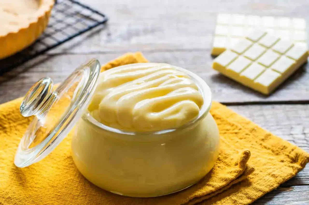 Un vasetto di cremosa crema al latte, con un coperchio di vetro appoggiato accanto. Sullo sfondo, una crostata e delle tavolette di cioccolato bianco.