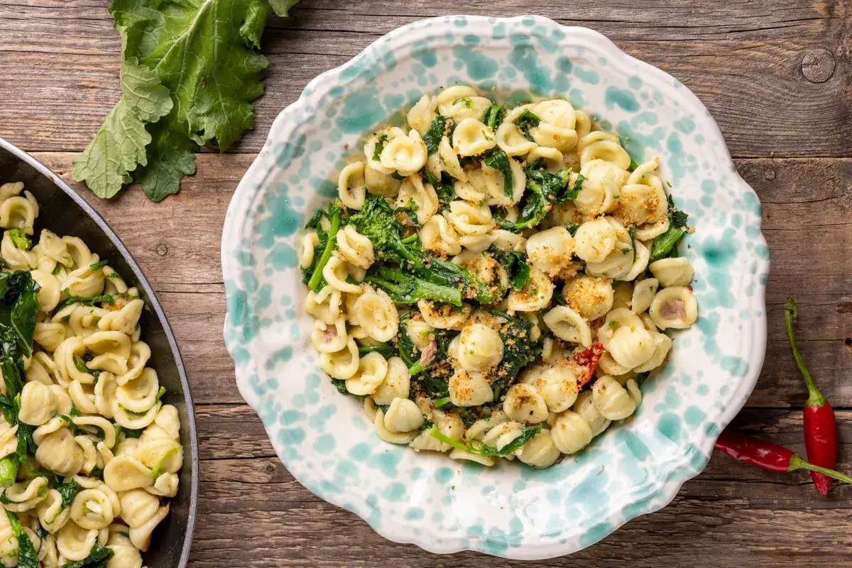 Piatto di orecchiette cime di rapa fumanti, condite con verdure verdi e pangrattato croccante, su un tavolo di legno rustico.