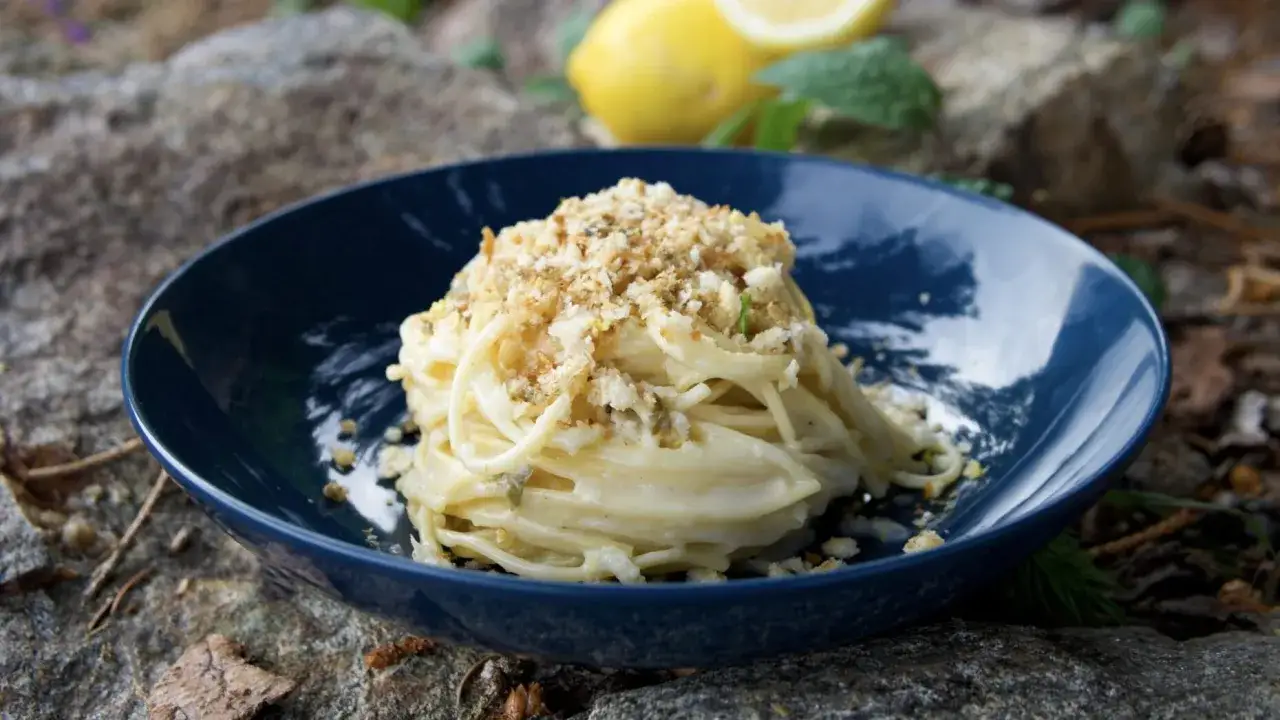 Un piatto di spaghetti al limone cremosi, guarniti con pangrattato croccante, su un piatto blu scuro, con limoni e menta sullo sfondo.