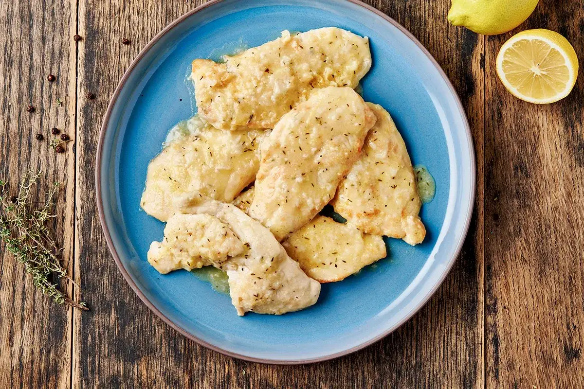 Piatto di pollo al limone con salsa cremosa e erbe aromatiche, guarnito con limone fresco e pepe nero su un tavolo di legno rustico.
